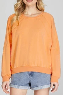 A woman wearing a tangerine orange French terry knit top with long sleeves and a round neckline, paired with denim shorts. Front view