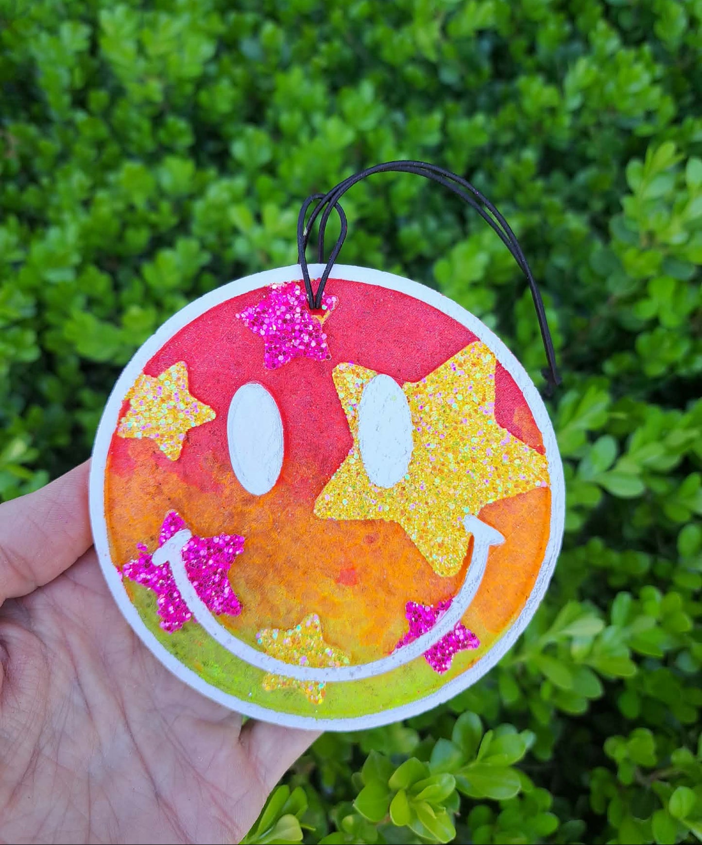 Circular freshie with star and smiley face design held against a green leafy background