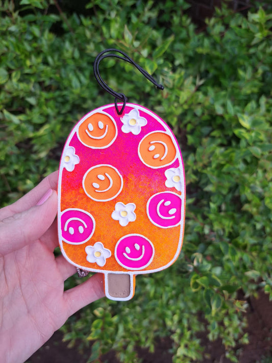A handmade, painted smiley face popsicle-shaped air freshener with ombre pink to orange color gradient, and decorative elements, attached with an elastic black string.