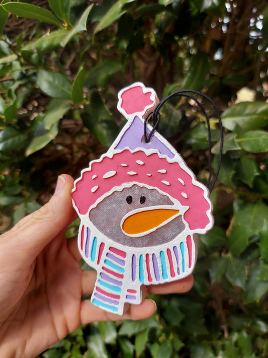 snowman in scarf freshie with black string held in hand. Green foilage background