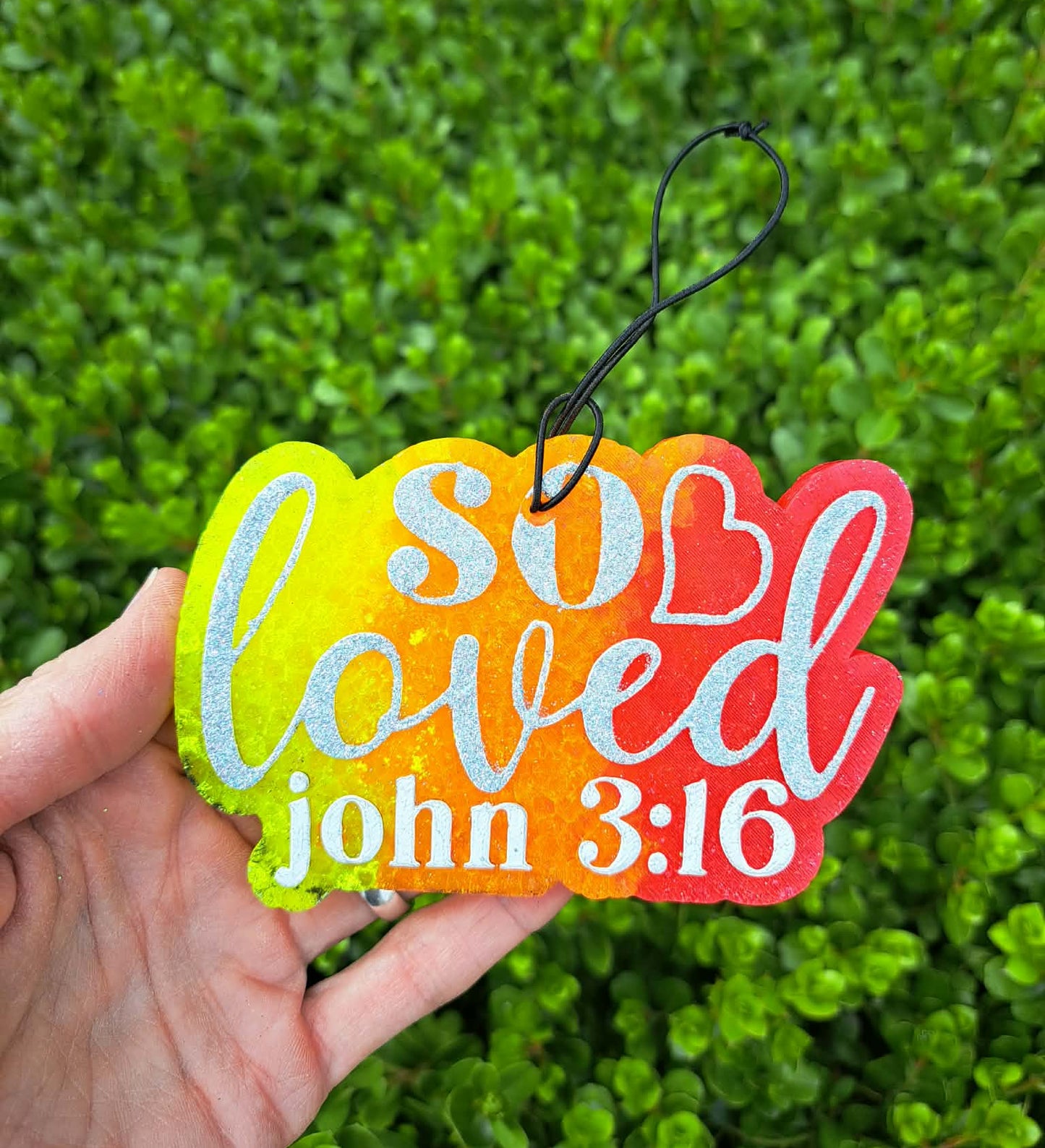 Colorful freshie with 'so loved' and 'John 3:16' text held against a green bush background.