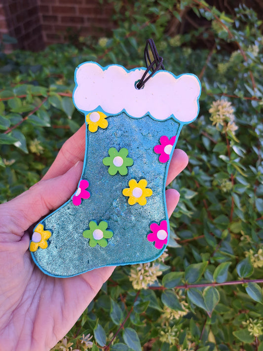 A hand holding a blue metallic air freshener shaped like a stocking with colorful flowers. Greenery background.
