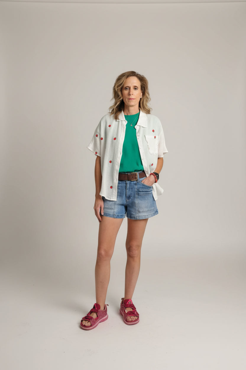 Woman standing in front of a blue curtain wearing a white jacket, green shirt, and denim shorts.