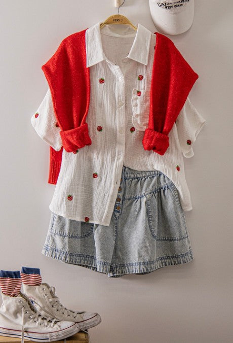Red cardigan over a white shirt with red buttons, denim shorts, and white sneakers on a wooden surface.