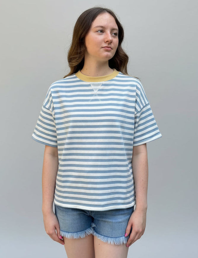 A young woman stands outdoors in front of a large green hedge and a stone pillar, wearing a light blue and white striped short-sleeve t-shirt with a yellow crew neck, paired with denim shorts.