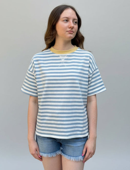 A young woman stands outdoors in front of a large green hedge and a stone pillar, wearing a light blue and white striped short-sleeve t-shirt with a yellow crew neck, paired with denim shorts.