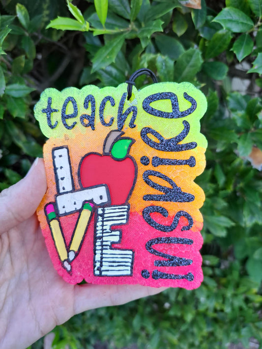 Colorful freshie with 'teach inspire' text held in front of green foliage