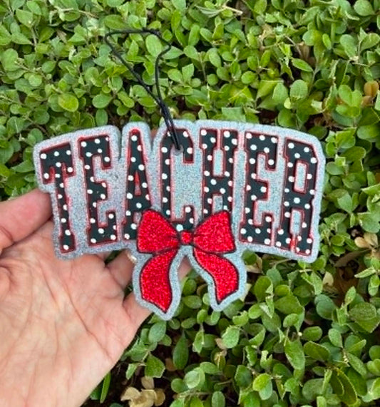 A hand holds a glittery 'TEACHER' word sign with a red bow, set against a background of green leafy bushes.