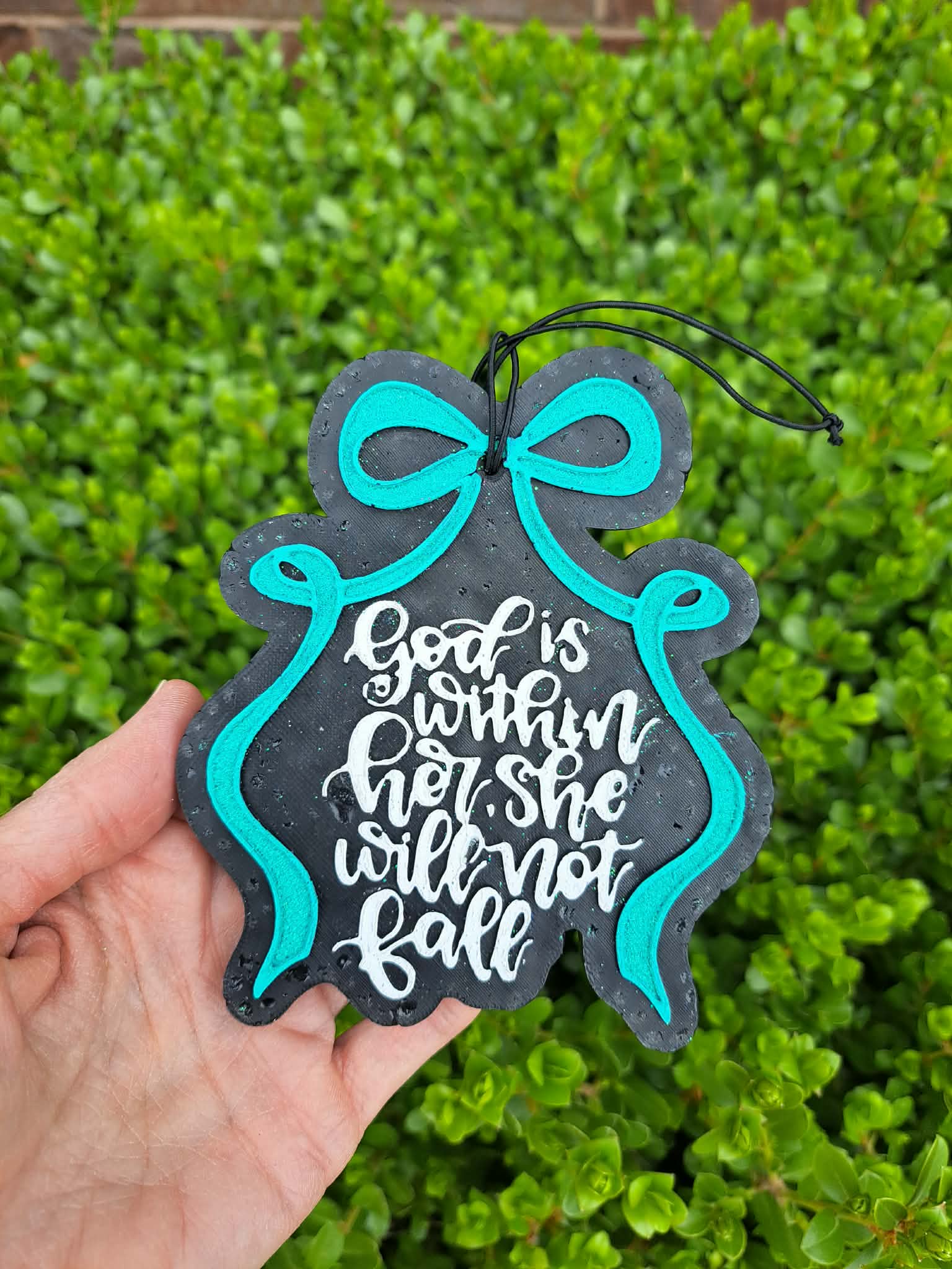 Decorative sign with 'God is within her, she will not fall' text held against a green bush background.