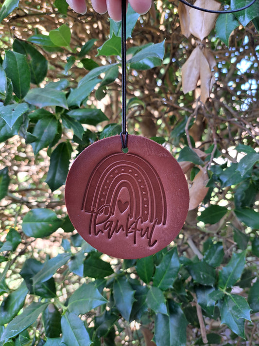 A brown leather air freshener with an embossed heart and the word 'Thankful' hanging on an elastic string against a leafy background.