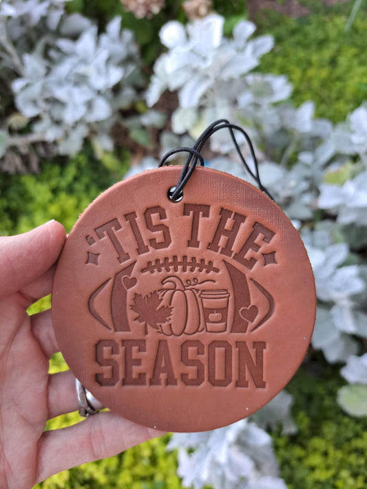 Round brown leather freshie with 'It is the Season' text held in front of greenery