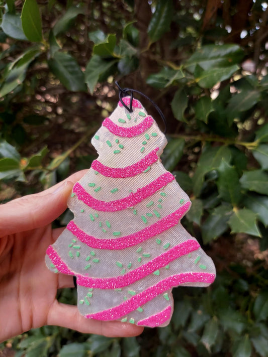 Hand holding a pink and green striped Christmas tree-shaped air freshener with white trimmings and a black elastic string. Greenery background