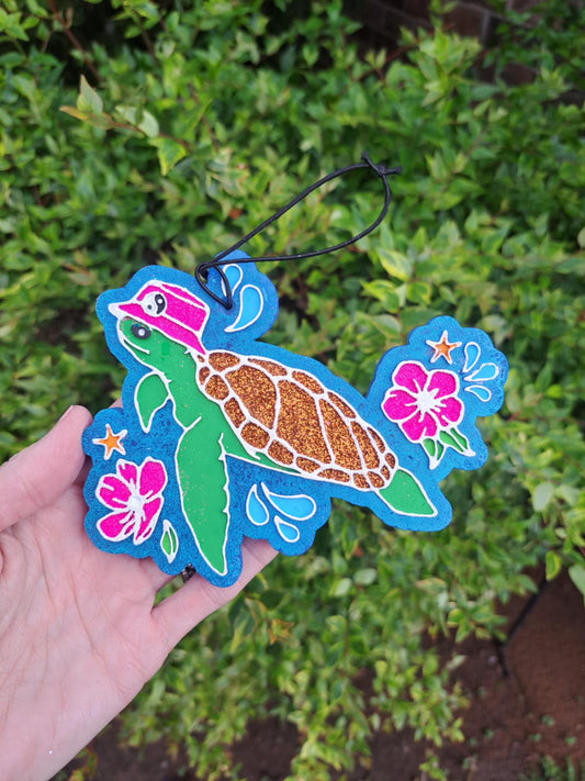 A handmade and painted hanging air freshener in the shape of a turtle with a floral pattern, predominantly in shades of blue and green, with a black elastic string for hanging.