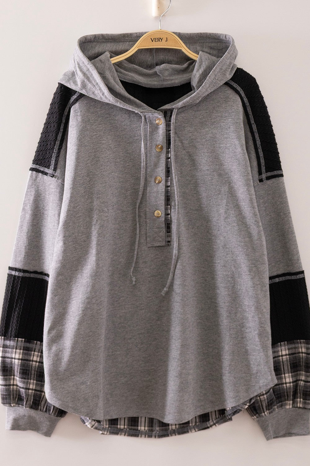 A gray hooded long-sleeve henley top with black knit shoulder panels and plaid cuffs, hanging on a wooden hanger against a plain light background.