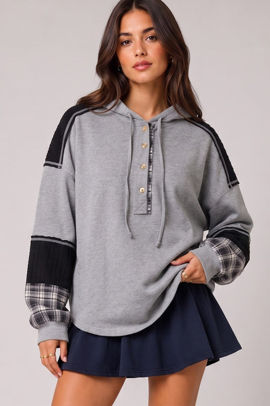 A woman modeling a gray hooded sweatshirt with black shoulder panels, a buttoned plaid collar, and plaid cuffs, paired with a navy pleated mini skirt against a light gray studio background.