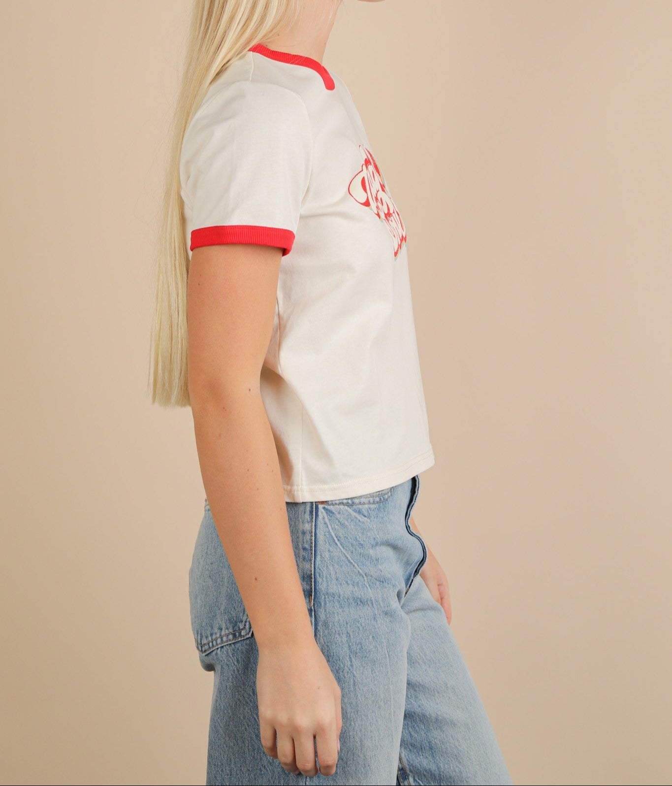Person wearing a white baseball-style shirt with red trim and blue jeans on a beige background
