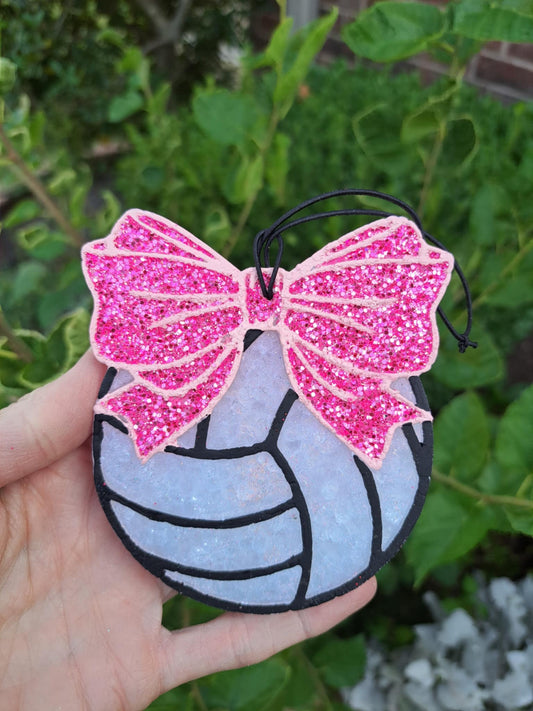 Volleyball air freshener with pink glittery bow held in a hand against a green leafy background.
