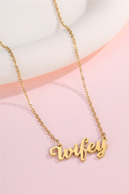 A gold chain necklace with a cursive script pendant spelling 'wifey', displayed on a soft pink and white background.
