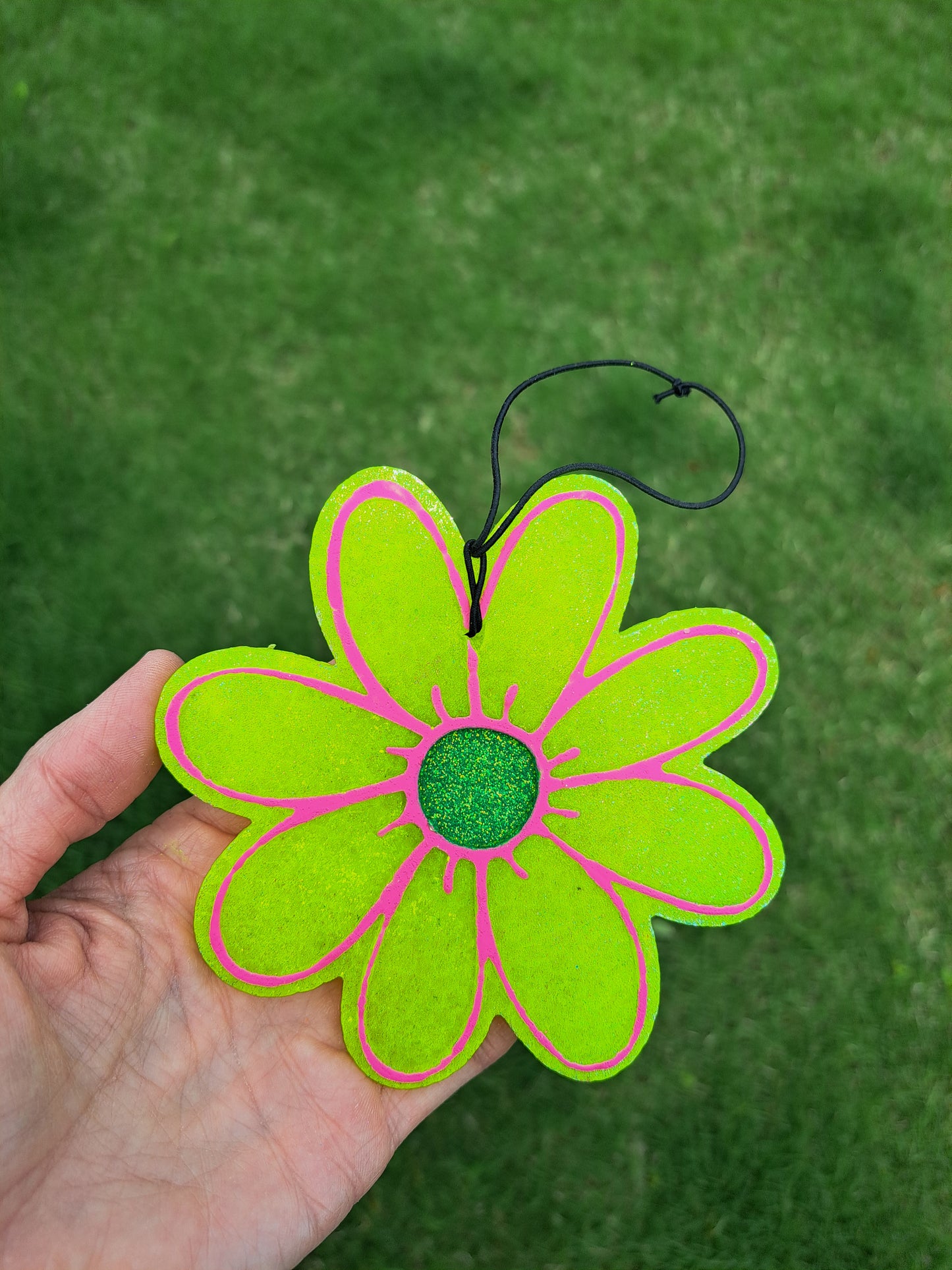 Hand holding a bright yellow daisy freshie with pink accents against a green background