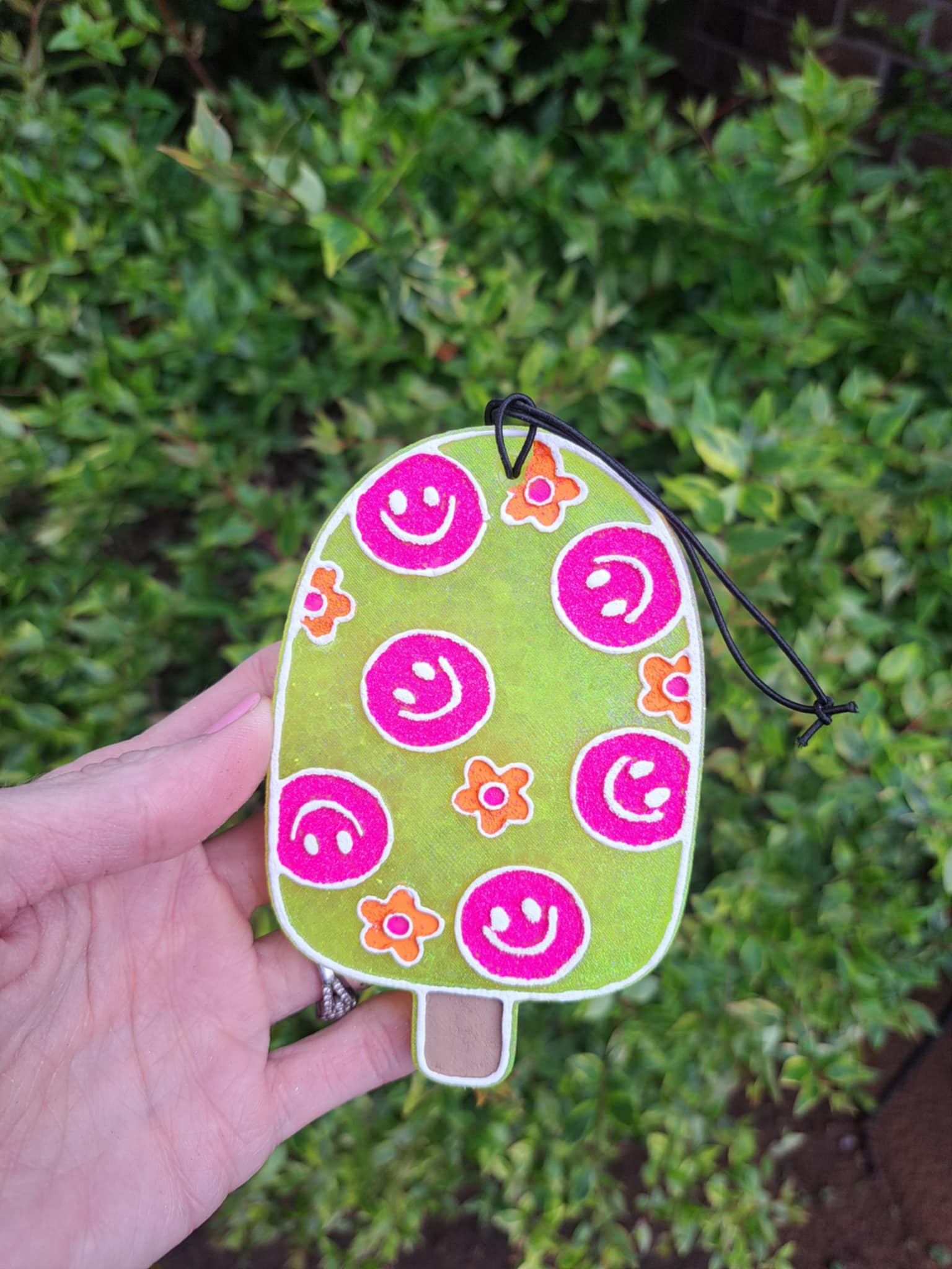 A handmade, yellow popsicle-shaped air freshener with a smiling face design and floral patterns, attached to an elastic black string.