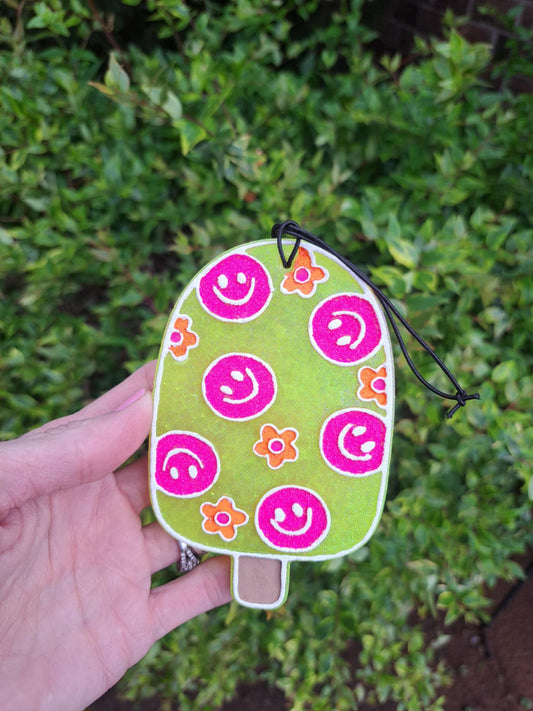A handmade, yellow popsicle-shaped air freshener with a smiling face design and floral patterns, attached to an elastic black string.