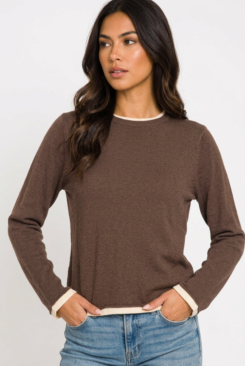 A woman wearing a brown long-sleeve knit top with contrasting cream ribbed trim at the crew neck and cuffs, paired with blue jeans, against a plain white background.