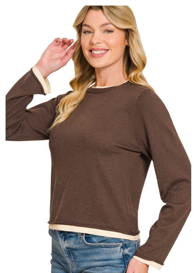 A woman wearing a brown long-sleeve knit top with contrasting cream ribbed cuffs and hem, paired with blue jeans, against a plain white background.