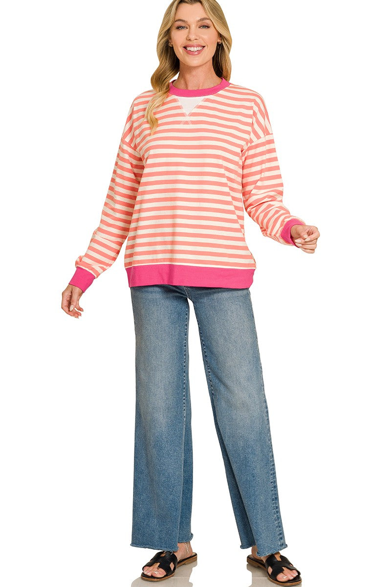 A woman modeling a long-sleeve crewneck sweater with horizontal peach and white stripes, featuring bright pink trim, paired with blue jeans and black sandals against a white background.