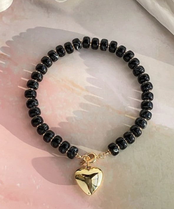 black beaded necklace with gold toggle clasp and dangling heart charm against a neutral background